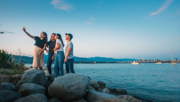 A group of young people taking a selfie on a rocky shoreline, with boats, mountains and the Vancouver skyline behind them