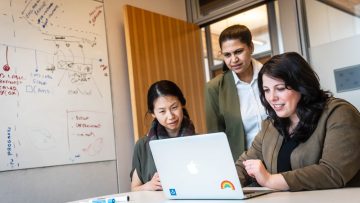 Three people grouped around a laptop screen in a meeting room