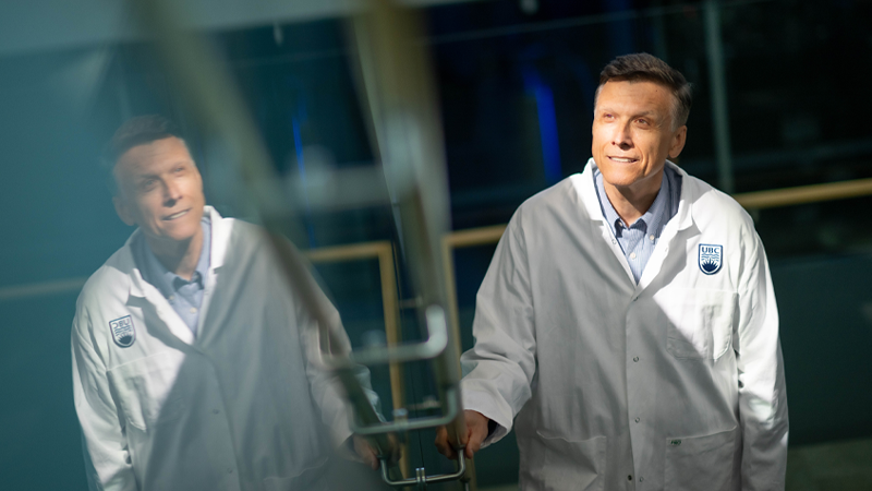 Dr. Erik Pioro smiling wearing a UBC lab coat. He is reflected in the mirrored wall next to him