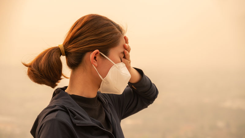 Person in smoke haze wearing a face mask, a hand held to their head